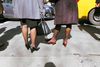 Rearview shot of 2 women with medium brown skin dressed in coats and heels standing on a city street and facing cars with shadows of other people nearby.