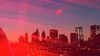 Hazy view of the New York City skyline at sunset with beams of red light stretching across in the foreground. 