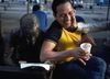 A man sitting by a statue of a man reading laughs as someone presses their hand into his face