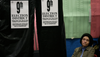 A woman wearing a knit hat sits next to a voting booth displaying signs indicating it is for voters in the 9th election district