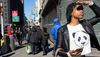 A woman wearing a shirt depicting a panda stands at the corner of a busy sidewalk, looking into the distance and holding her hand above her brow to block the sun