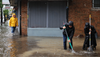 In the right of frame, two people in black raincoats dig beneath floodwater up to their calves with shovels; in the left of frame, a man in a yellow raincoat approaches