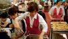 A waitress at a restaurant reaches for a dish for a group of customers sitting to the left of frame from a rolling cart; another waiter rolls a cart forward in the right of frame behind her