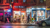 Storefronts on Essex Street in New York City's Chinatown are lit up at night as two people walk by, one with a bike and one with an umbrella