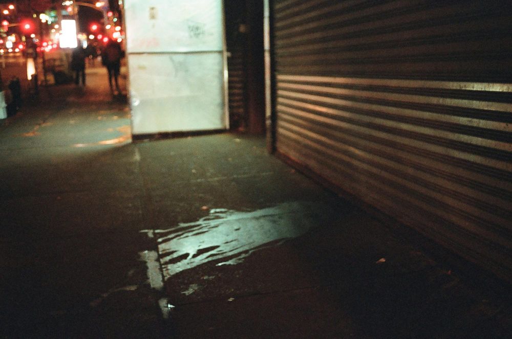 Nighttime shot of a city sidewalk with street lights, traffic, and people walking in the distance. On the right is a closed pull-down, corrugated storefront door next to a dirty sidewalk.