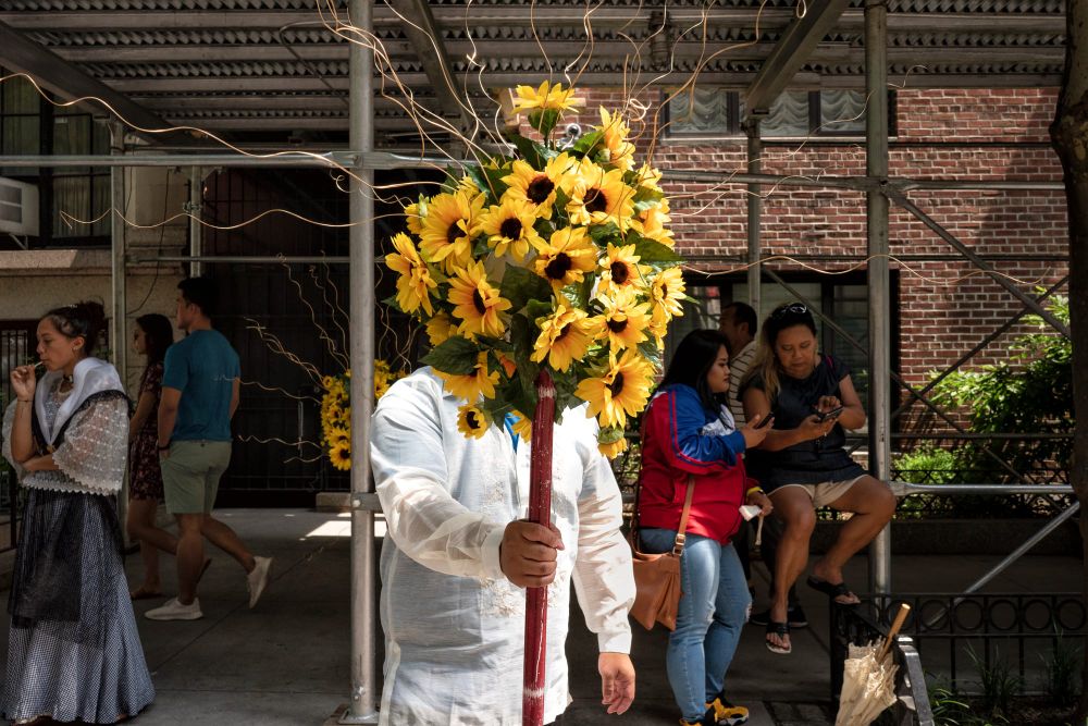 A man standing below scaffolding holds out a large bouquet of sunflowers to the camera as people walk behind him