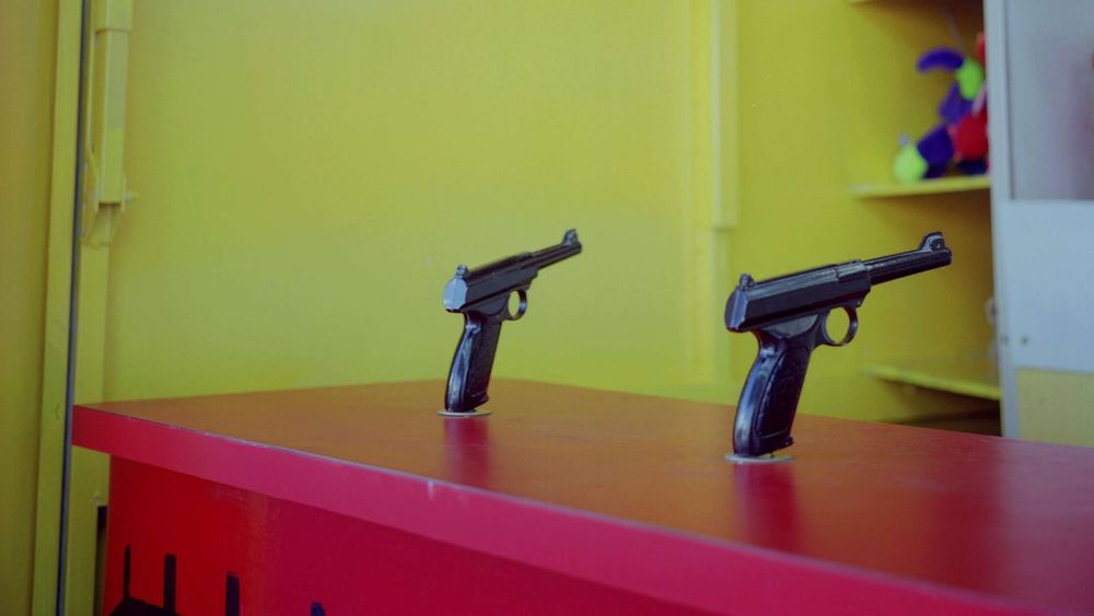 Closeup of a carnival table with fixed toy pistols aimed toward a wall. Colorful prizes appear on a shelf in the upper left.
