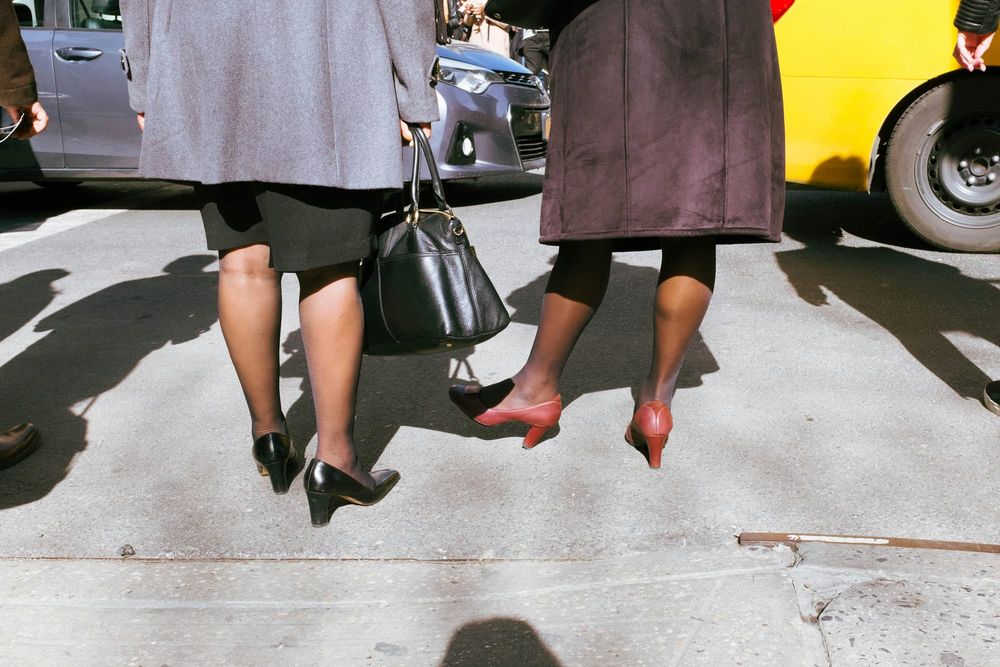 Rearview shot of 2 women with medium brown skin dressed in coats and heels standing on a city street and facing cars with shadows of other people nearby.