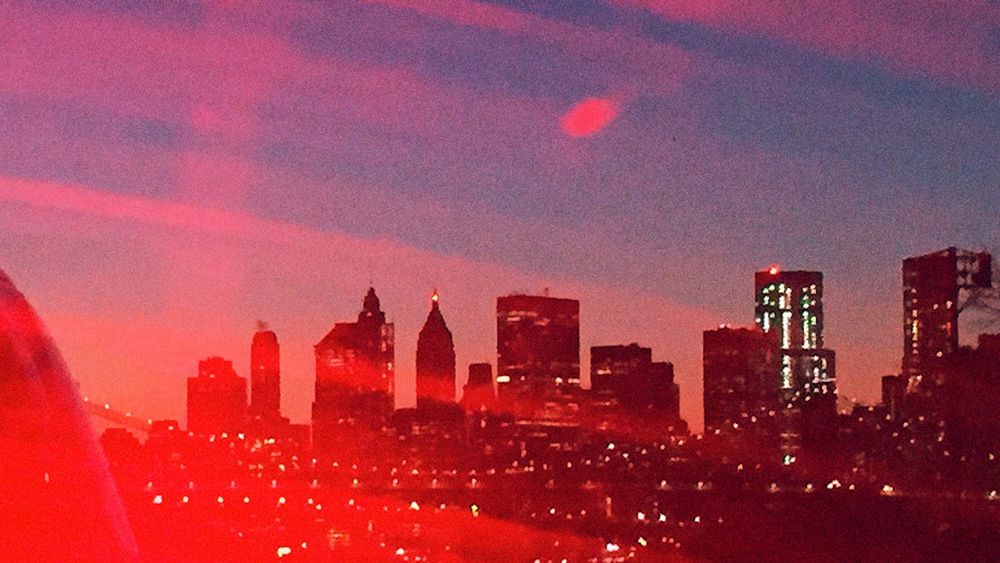 Hazy view of the New York City skyline at sunset with beams of red light stretching across in the foreground.