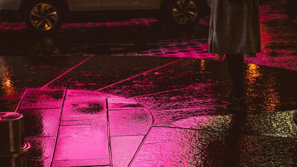 Nighttime closeup of a wet city sidewalk with a standing figure wearing boots and a coat facing out towards a street with a passing car.