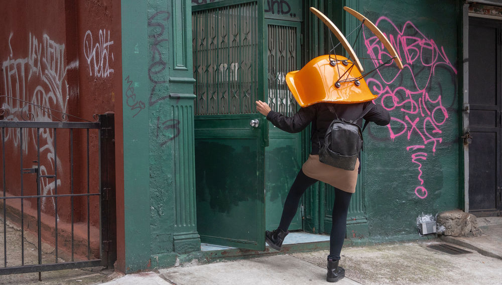 A woman opens the door to her apartment building while carrying an orange plastic chair on top of her head