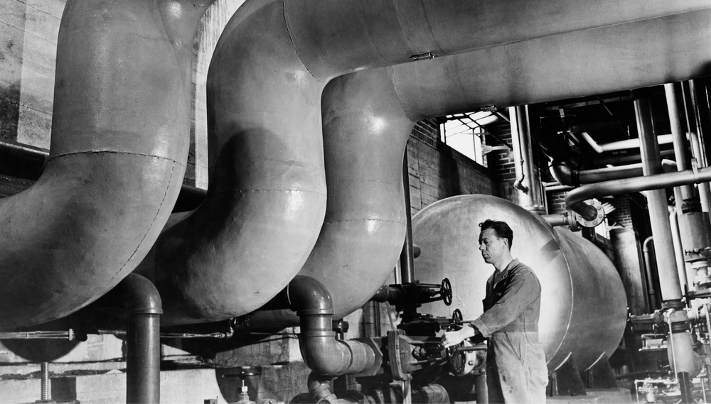 A black and white image of a technician operating an old-fashioned steam boiler in a New York City apartment basement