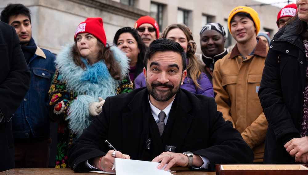 Mamdani signs an executive order during a news conference at Grand Army Plaza with a row of supporters standing behind him