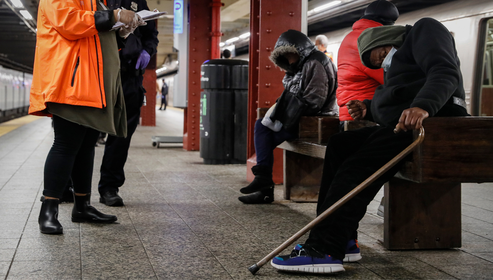 A police officer and a homeless outreach worker assist a homeless man sleeping on a bench on a Subway platform