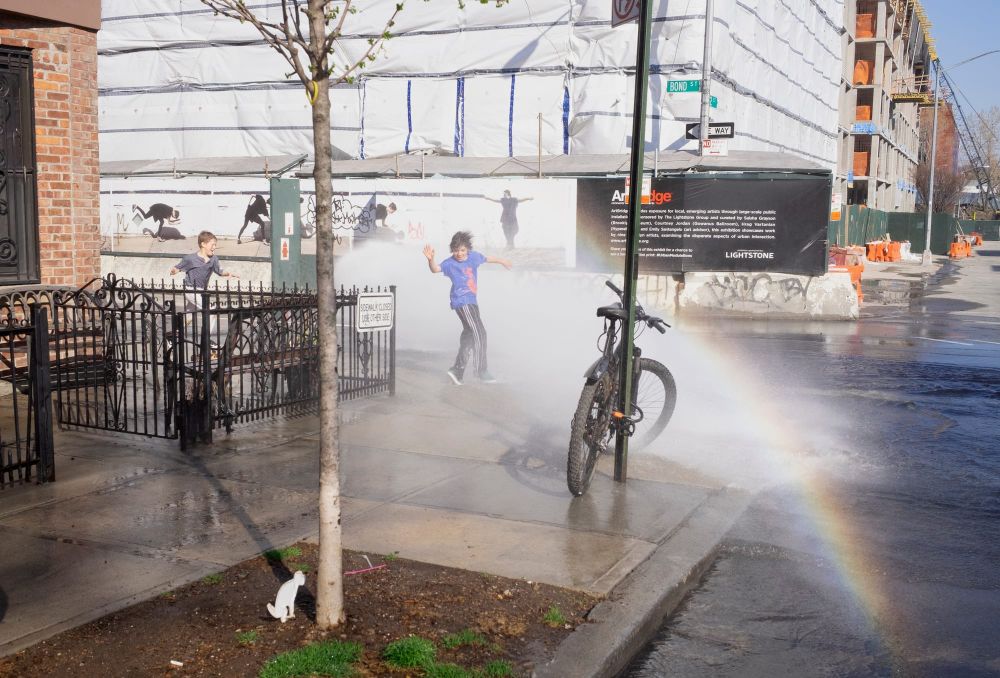 The light reflects a rainbow as kids play in a sprinkler on a corner near a construction site