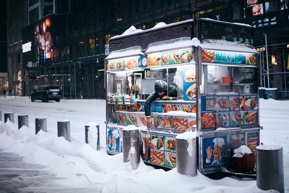 A Halal cart vendor reaches out of his cart during a snowstorm