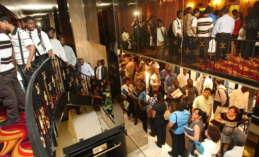 People line up on and below the stairs leading to a hiring fair; next to the stairs is a wall of mirrors that reflects some of their images back