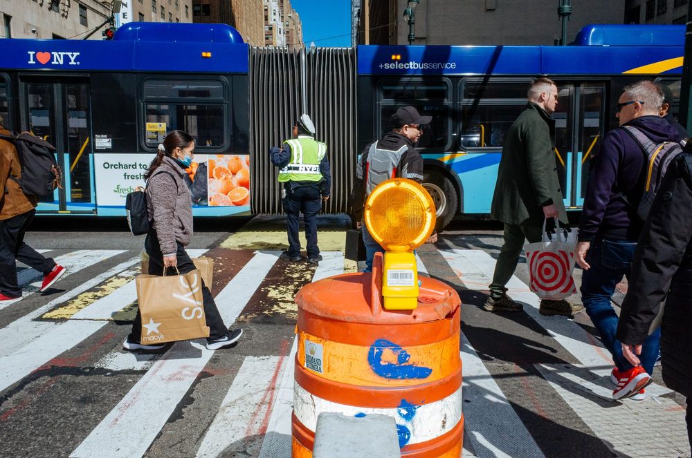 People cross a New York City crosswalk as a bus passes by behind them