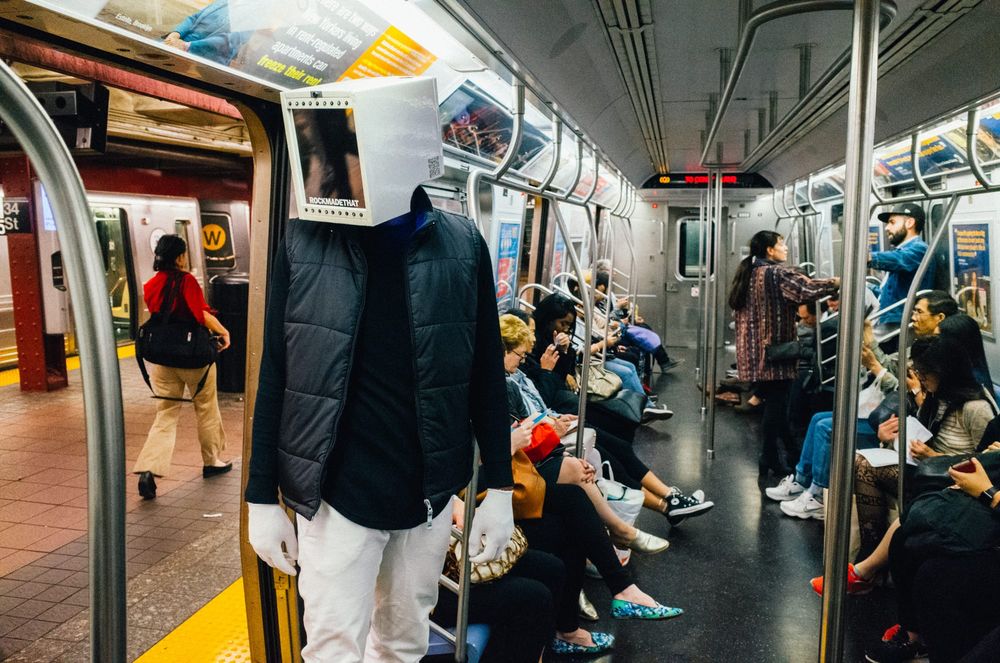 A man wearing a mask shaped like a computer over his head stands on a subway train