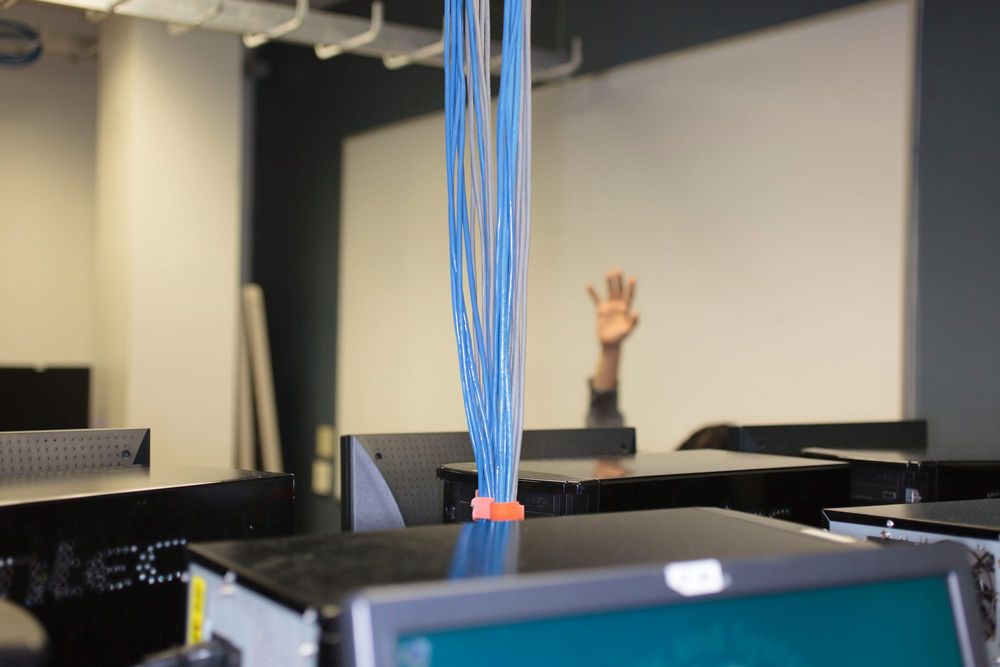 A person raides their hand above a computer in a room full of computers where blue wires hang down from the ceiling