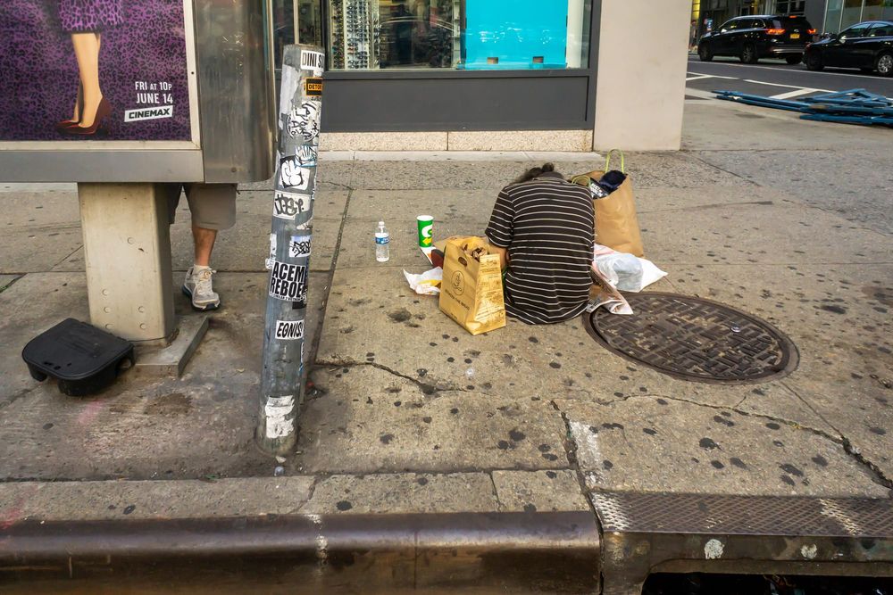 A homeless man sits on the sidewalk next to a man standing behind an advertising board