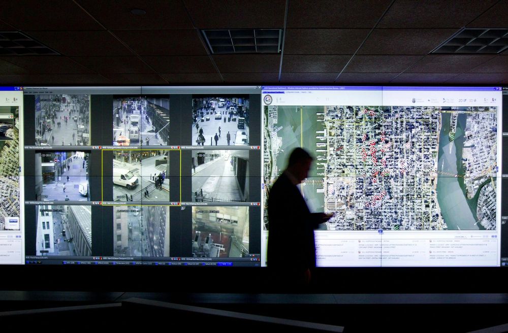 A man walks past a wall of surveillance camera feeds and a map