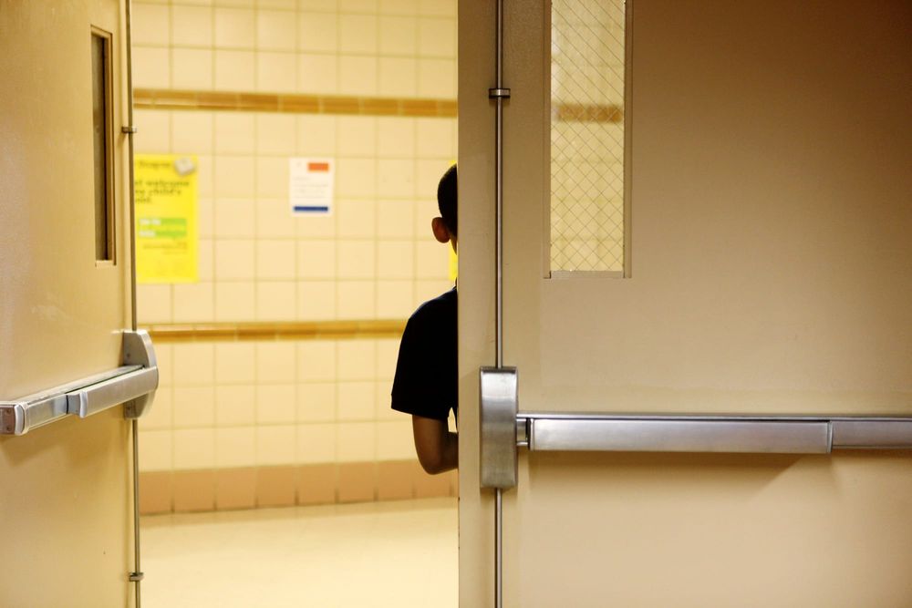 An elementary school boy stands behind a door at his school with his back to the camera; half of his body is obscured by the door, half is visible