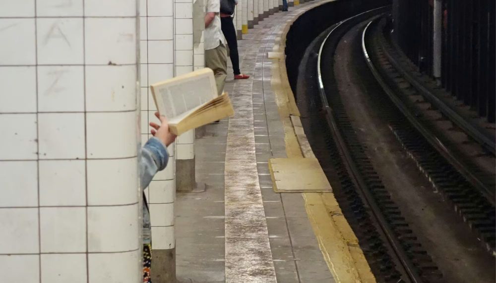 A hand holding a book is visible behind a subway platform column