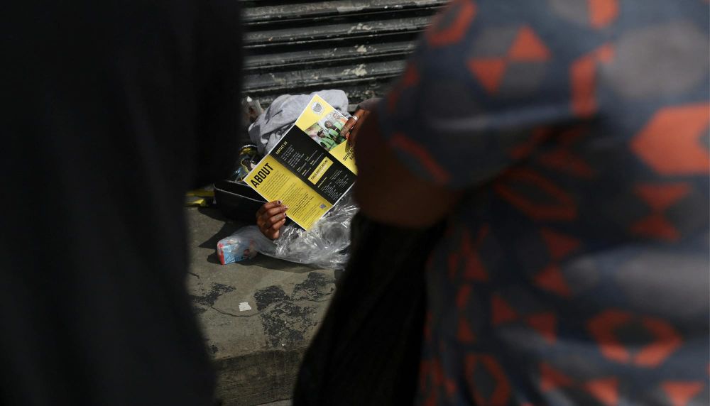 A person lying on the sidewalk reads literature given to them by Outreach Public and Safety team members from the overdose prevention center OnPoint
