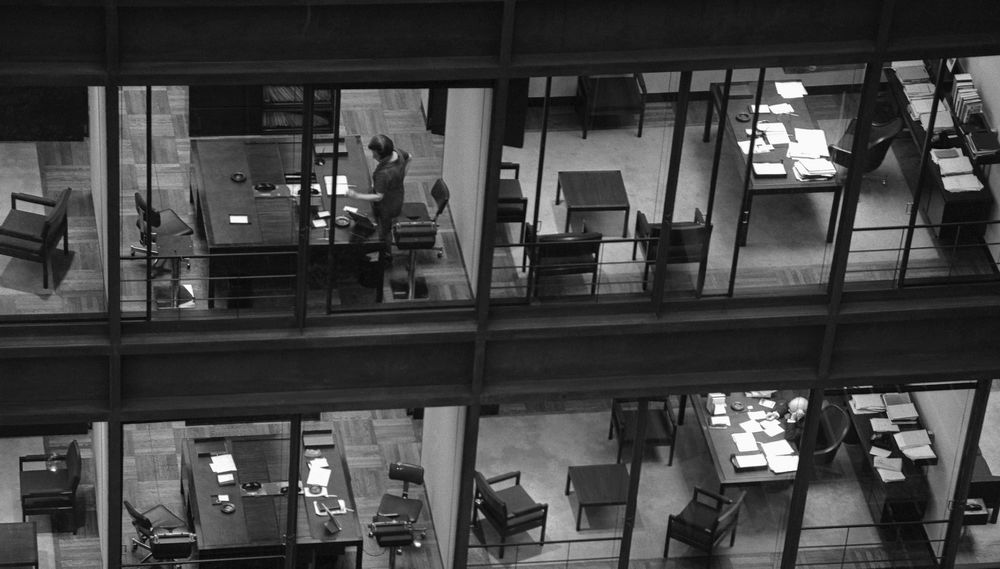 A row of windows in an office building, through which you can see the people working inside; shot in black and white