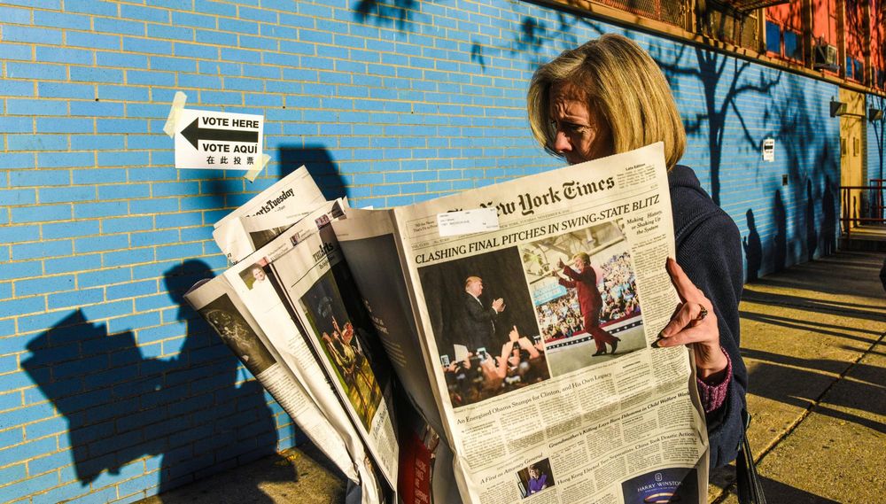 A woman reads a copy of the New York Times outside of a building with a blue wall; on the wall is taped a sign with the words "Vote Here" printed underneath