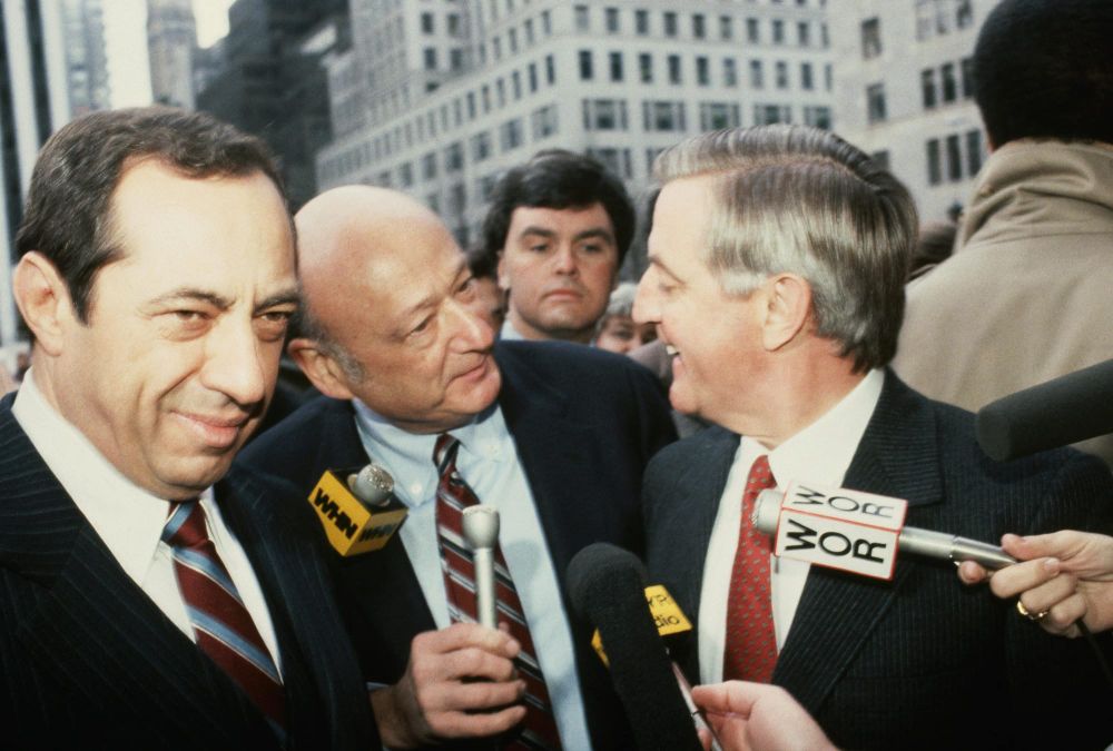 Three men in suits are interviewed by reporters holding microphones