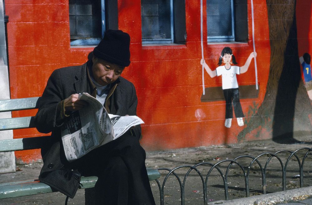 An older man reads a newspaper on a bench by a mural