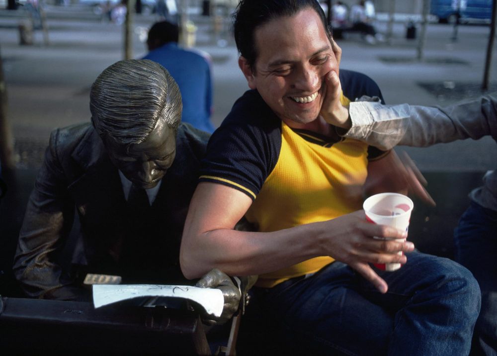 A man sitting by a statue of a man reading laughs as someone presses their hand into his face