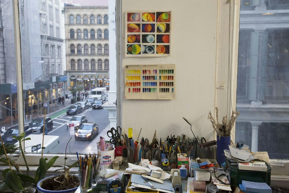 A desk covered in art supplies in front of a window looking out onto a Manhattan street