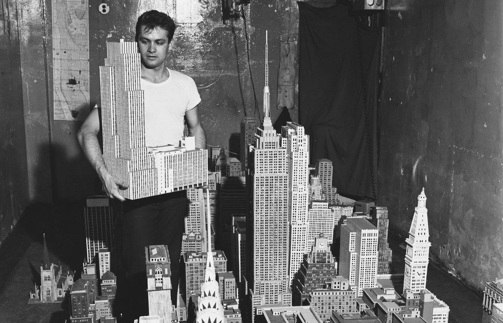 A man creates a miniature scale model of New York City, holding up one of the buildings