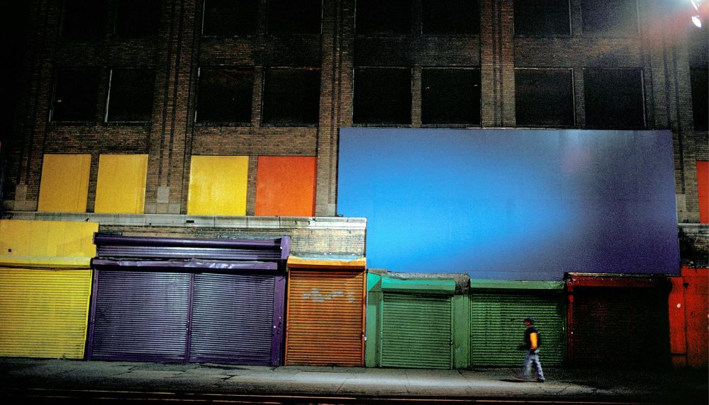 A man walks in front of a row of buildings with some windows boarded up with colorful boards and their doors painted a variety of bright colors