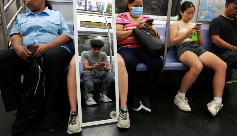 People sit on the subway with a full length mirror reflecting a person opposite them