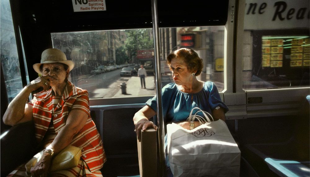 Two elderly women sit on a bus with large bags