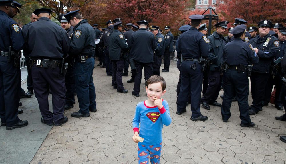 A young boy in superman pajamas stands in front of a crowd of NYPD offices, who are all facing away from him