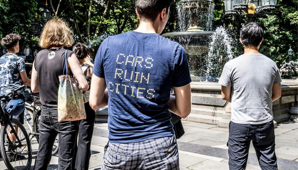 A person stands with their back to the camera in a blue t-shirt with writing on the back in yellow saying "Cars ruin cities"