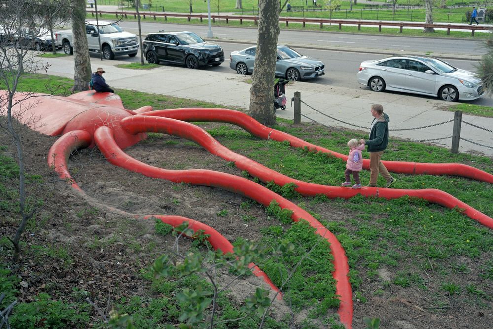 A toddler's father walks with her next to a large sculpture of a squid at a park to meet her mother on the other side