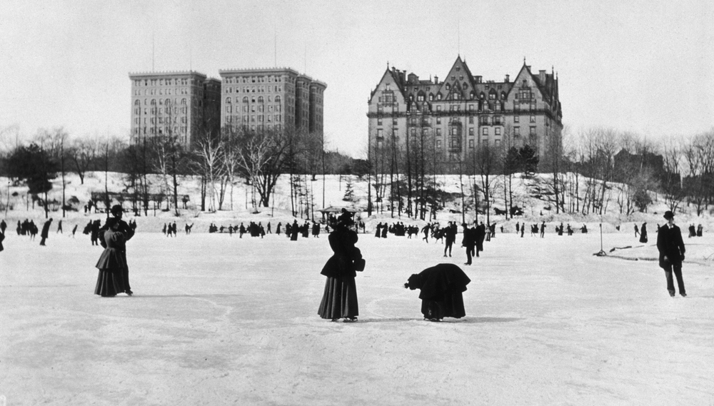People ice skate on a frozen pond in Central Park, buildings behind