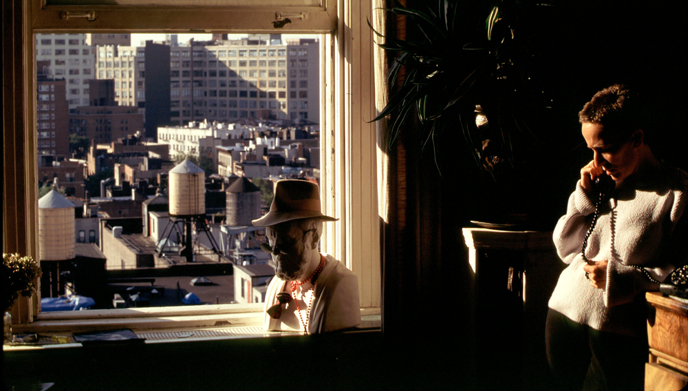 A person stands next to a window in their apartment taking a phone call; outside the window can be seen water tanks on rooftops