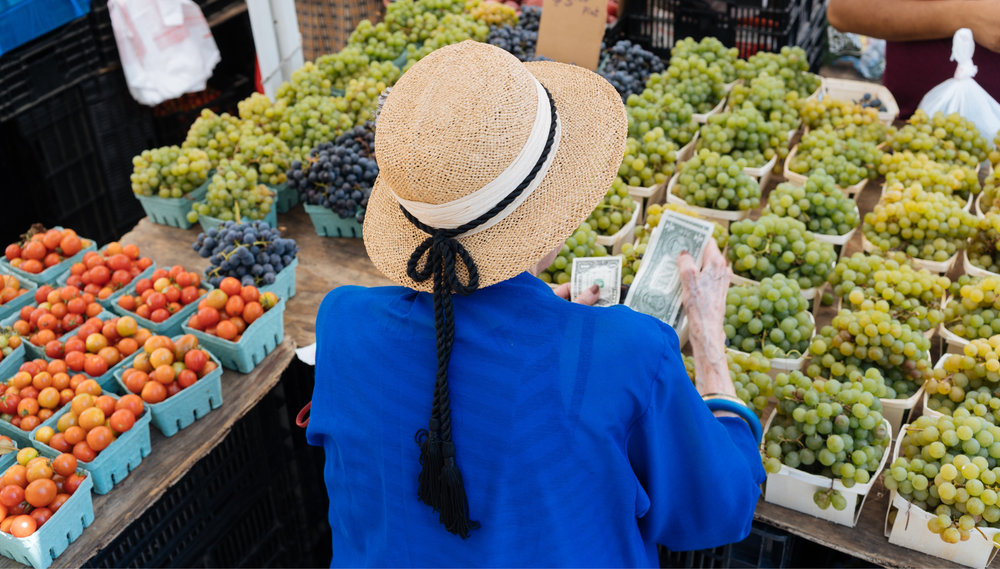 A woman takes out cash in front of a table of grapes and cherry tomatoes at a grocery market