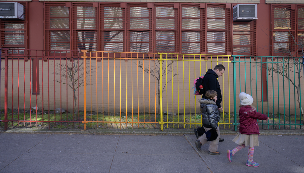 A father and two young children walk past a school with a gate painted in rainbow colors