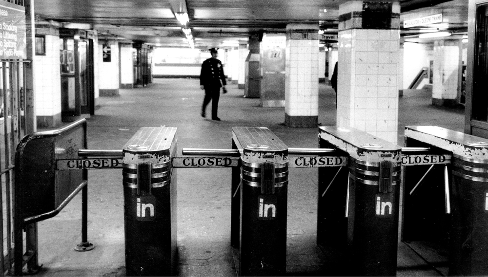 A black and white image of a police officer walking in a subway station behind a row of closed turnstiles