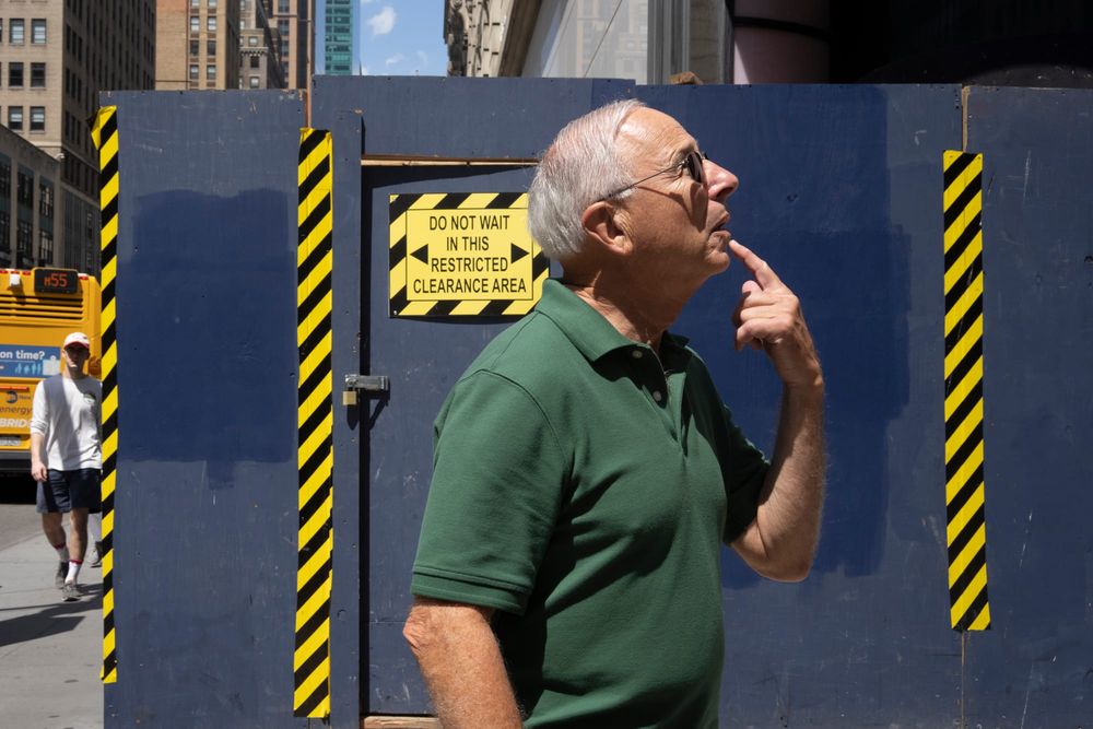 A man standing in front of a construction site holds his index finger to his chin and looks upward