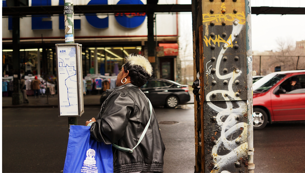 A woman wearing a leather jacket looks at the bus schedule at a bus stop along East Flatbush Ave in Brooklyn