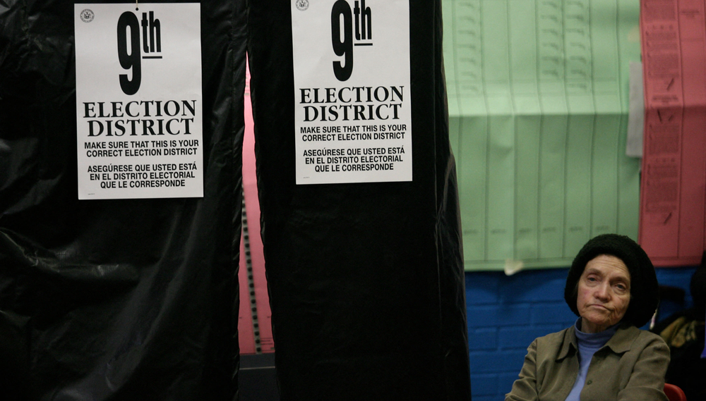 A woman wearing a knit hat sits next to a voting booth displaying signs indicating it is for voters in the 9th election district